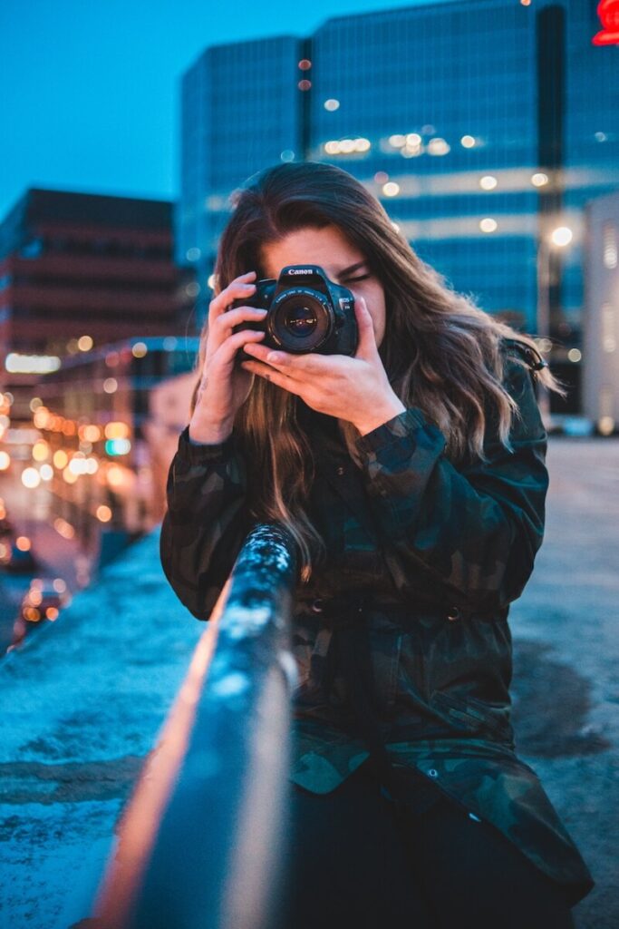 A young woman with long hair, partially obscuring her face, is taking a photo with a Canon DSLR camera. She is outdoors on a rooftop at dusk, with city buildings illuminated in the background.
