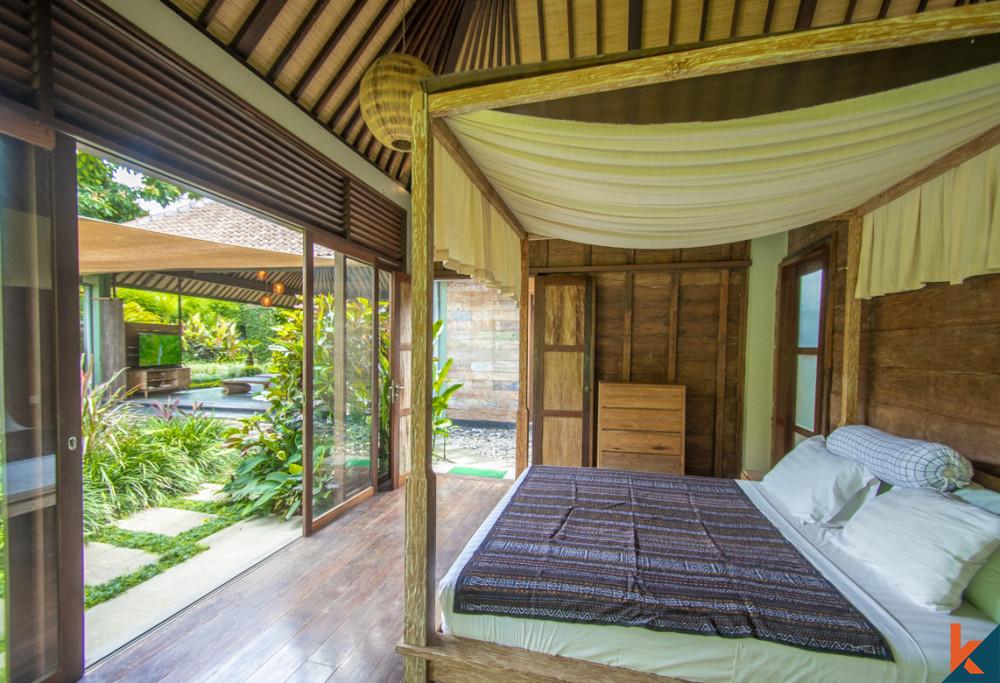 A cozy bedroom in an Ubud villa with a panoramic view of the garden, featuring traditional Balinese woodwork and a canopy bed, blending comfort with natural beauty.